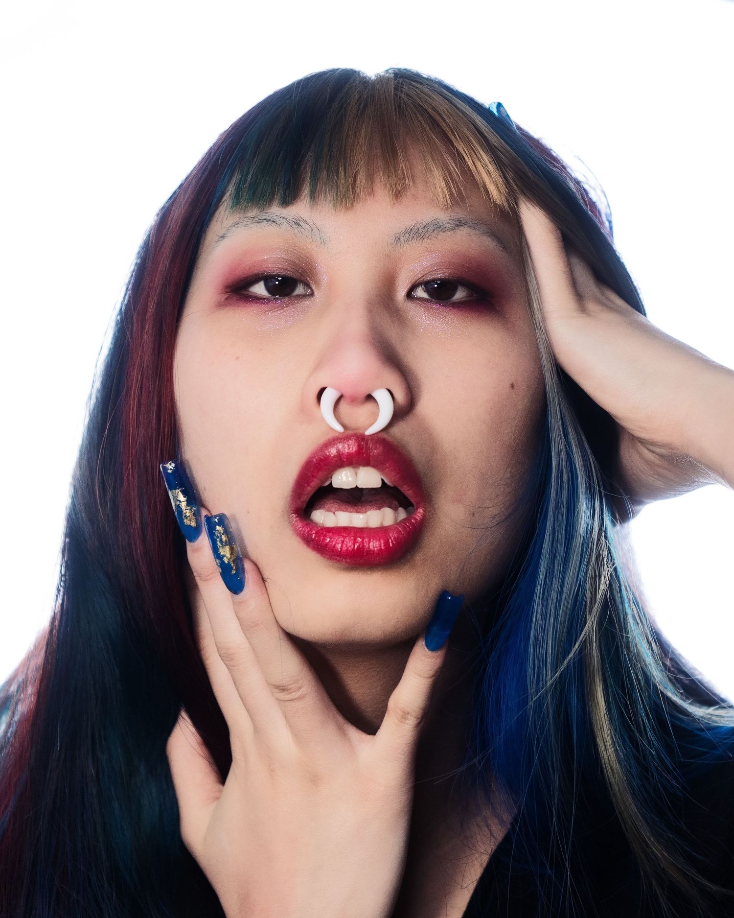 Young woman with colorful hair, septum piercing, dramatic red makeup, and decorative blue nails.