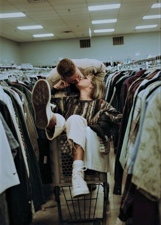 Couple sharing a romantic kiss in a thrift store shopping cart surrounded by racks of vintage clothing.