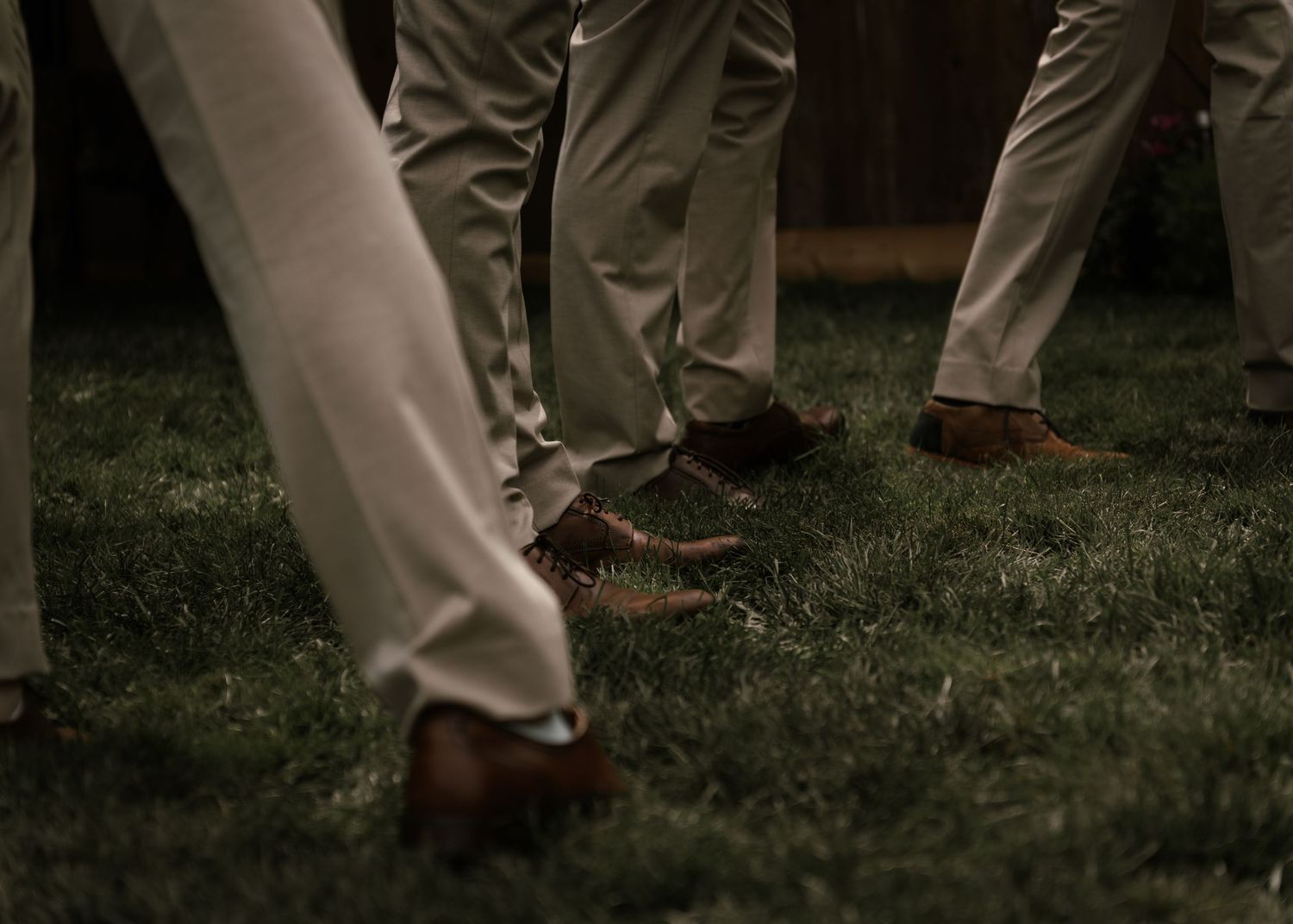 Men in khaki pants and brown dress shoes standing on green grass at an outdoor event.