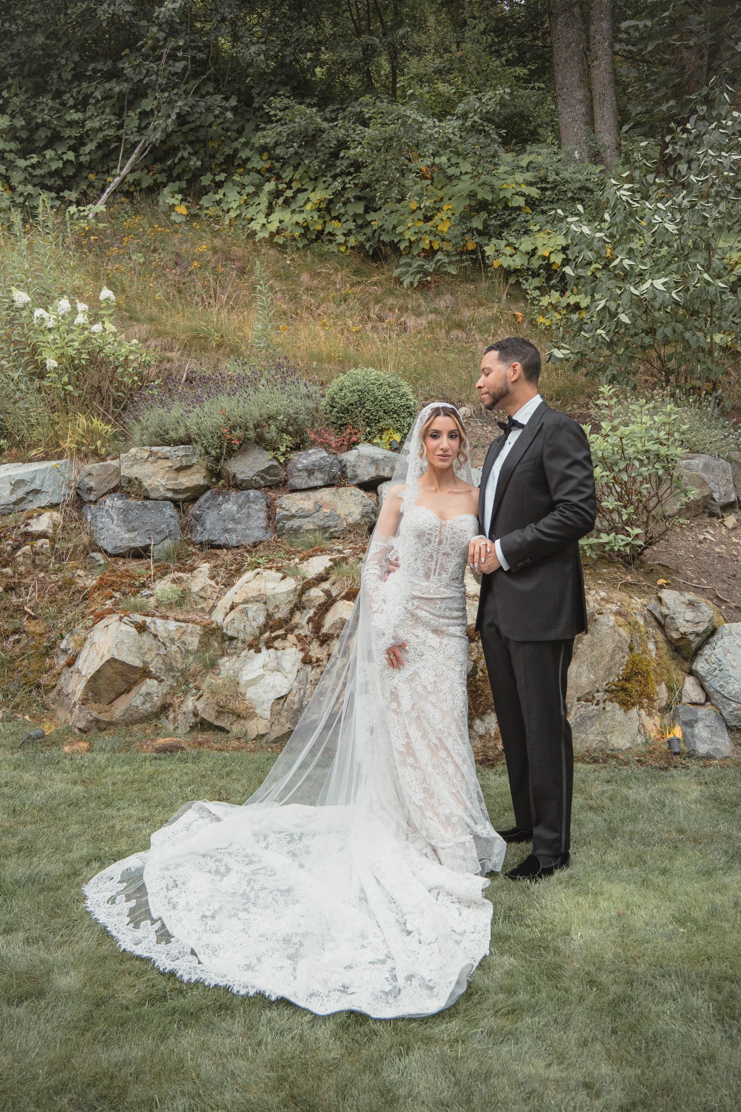 Bride in lace gown with cathedral veil stands with groom in tuxedo on garden lawn with stone wall backdrop.