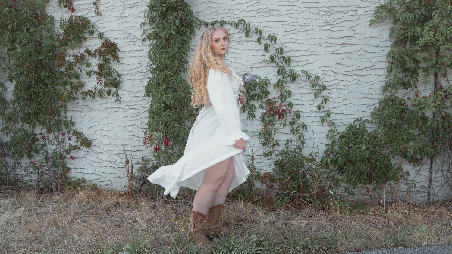 Blonde woman in flowing white dress twirls near ivy-covered white brick wall outdoors.