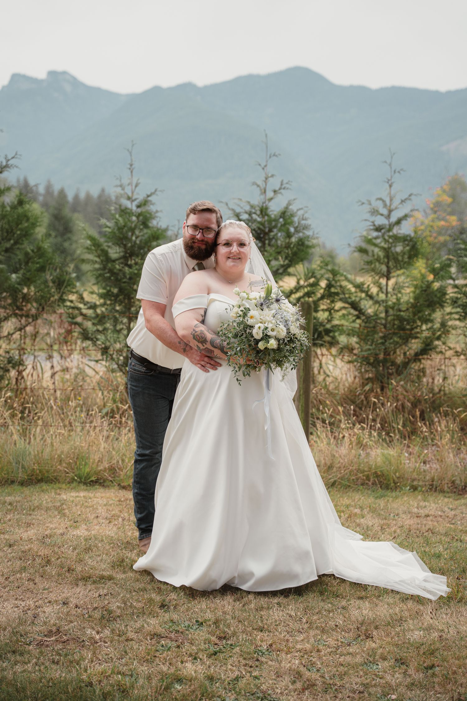 Bride and groom posing outdoors in a mountain meadow, bride in white gown holding floral bouquet.