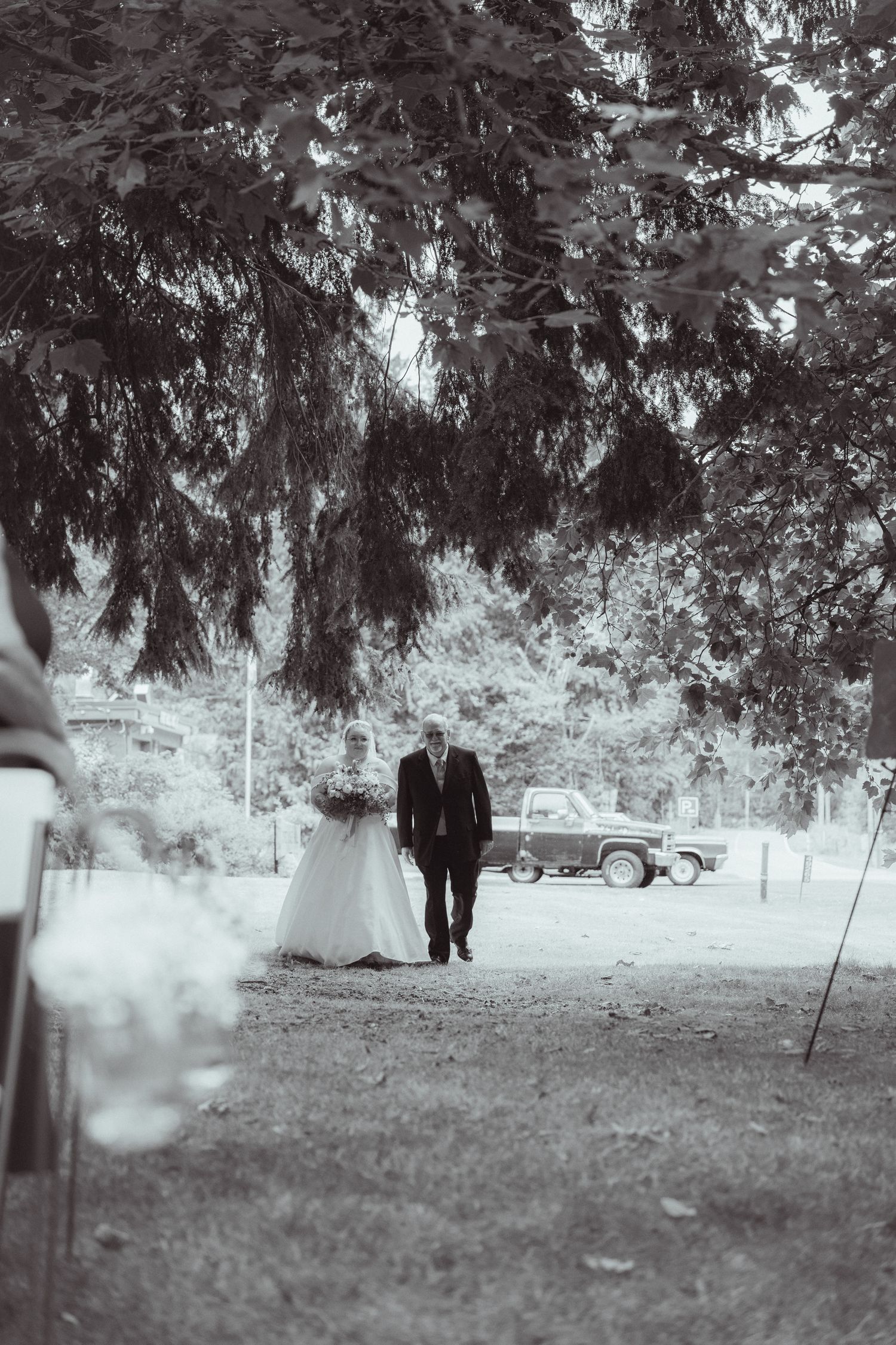 Black and white photo of bride and escort walking toward outdoor wedding ceremony aisle.