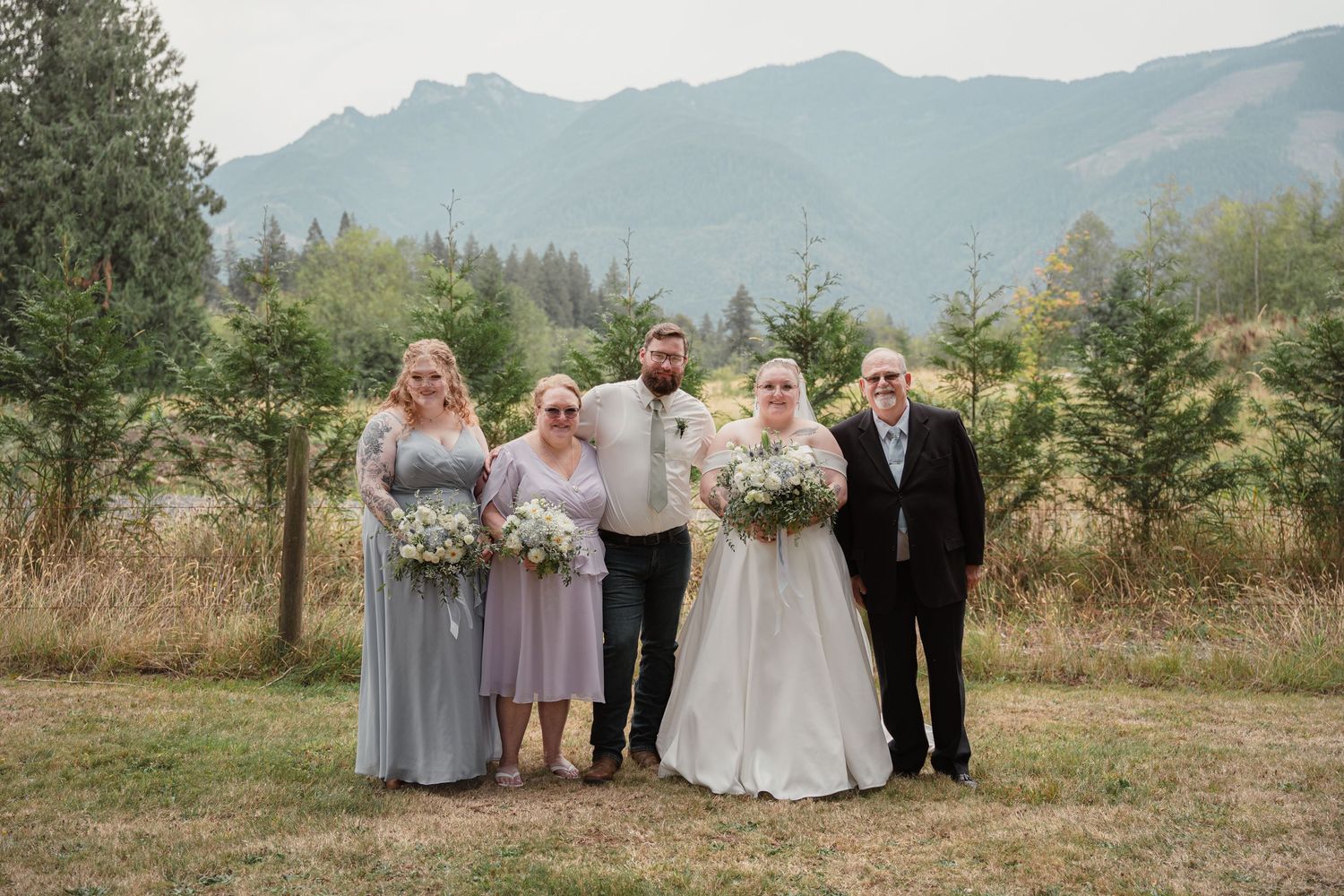 Wedding party poses outdoors with mountain backdrop, bride in white gown flanked by bridesmaids in blue dresses.