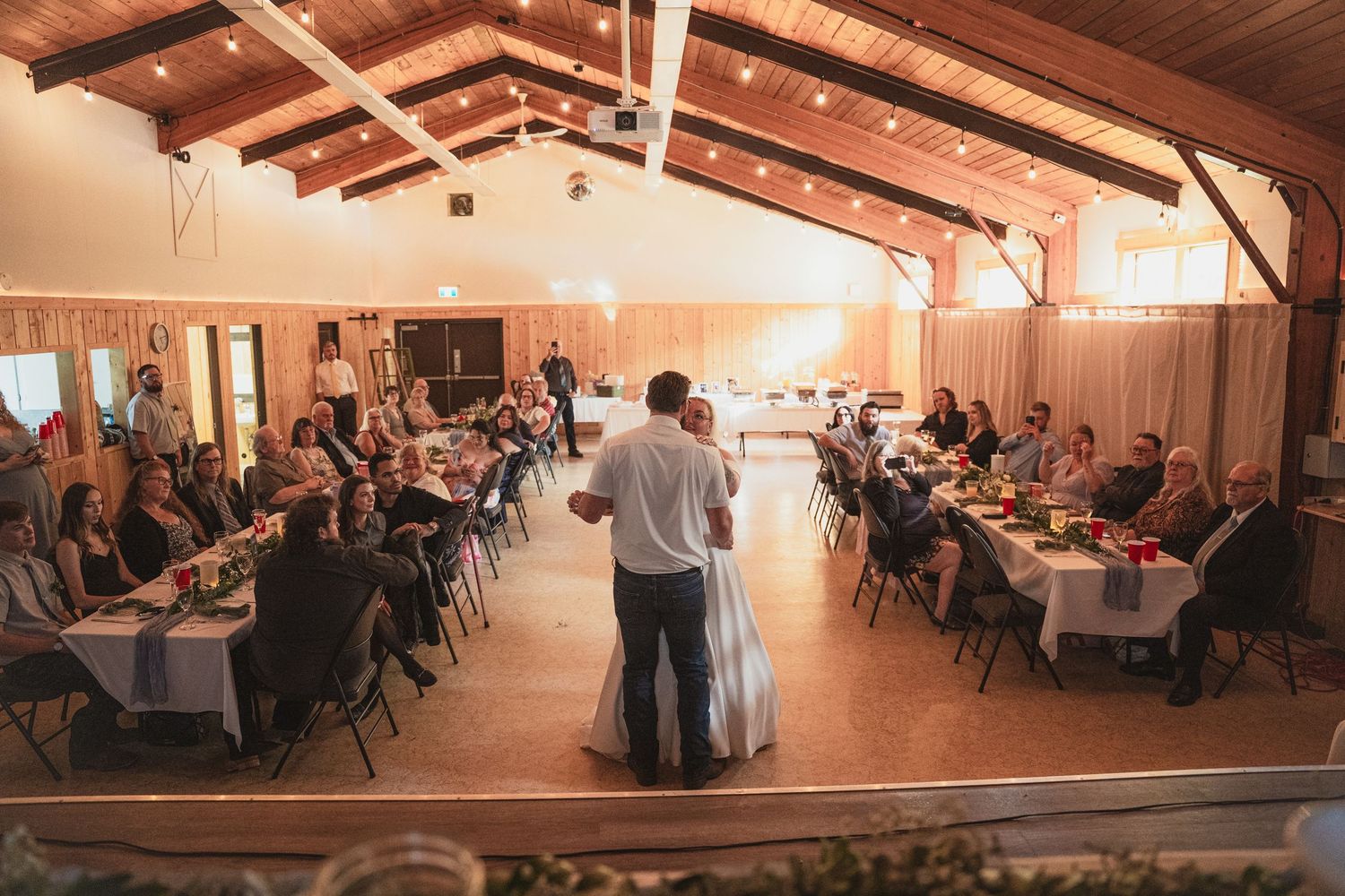 Aerial view of wedding reception in rustic barn venue with guests seated at long tables as couple shares first dance.