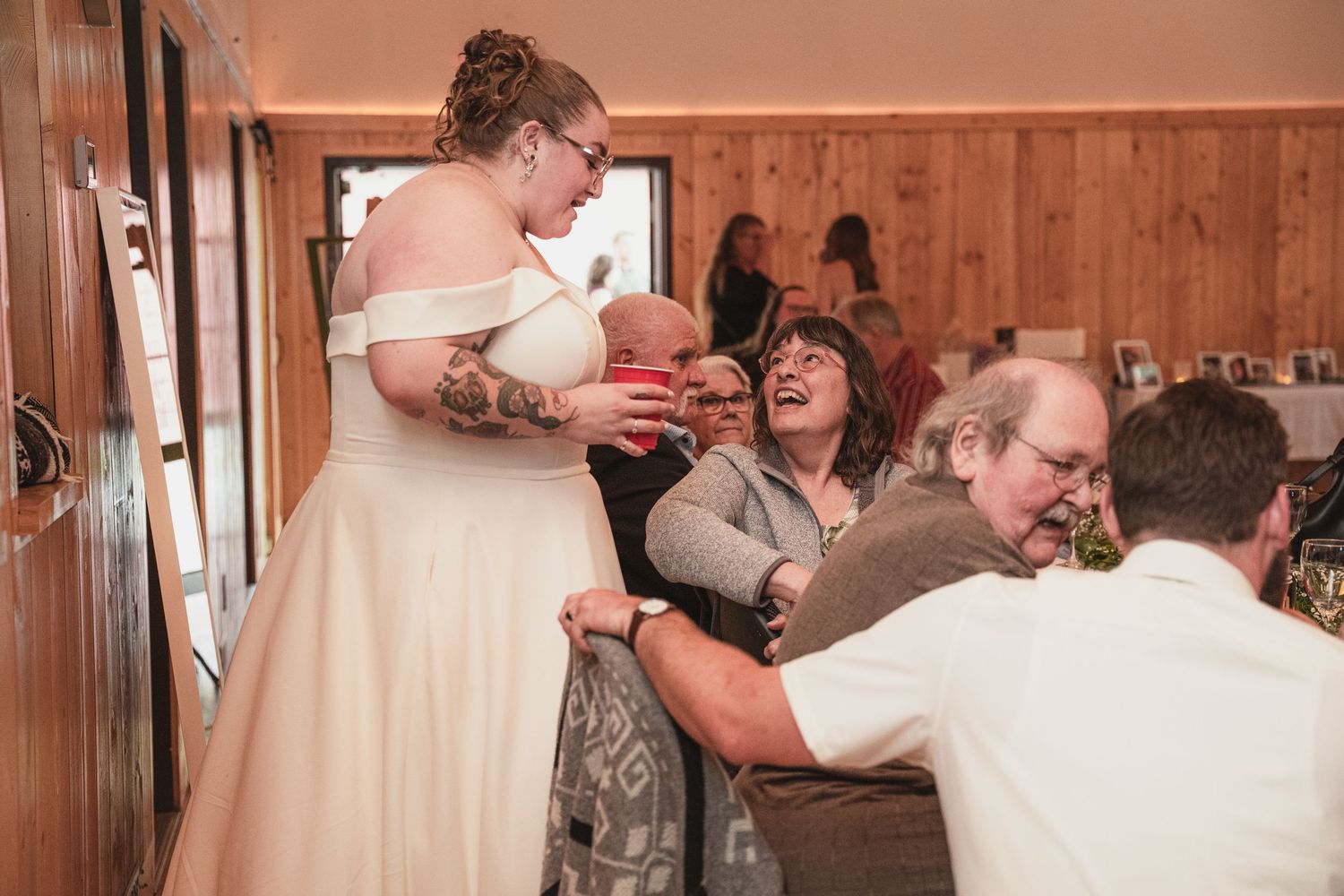 Bride in white off-shoulder gown sharing emotional hug with family members at rustic wedding reception.