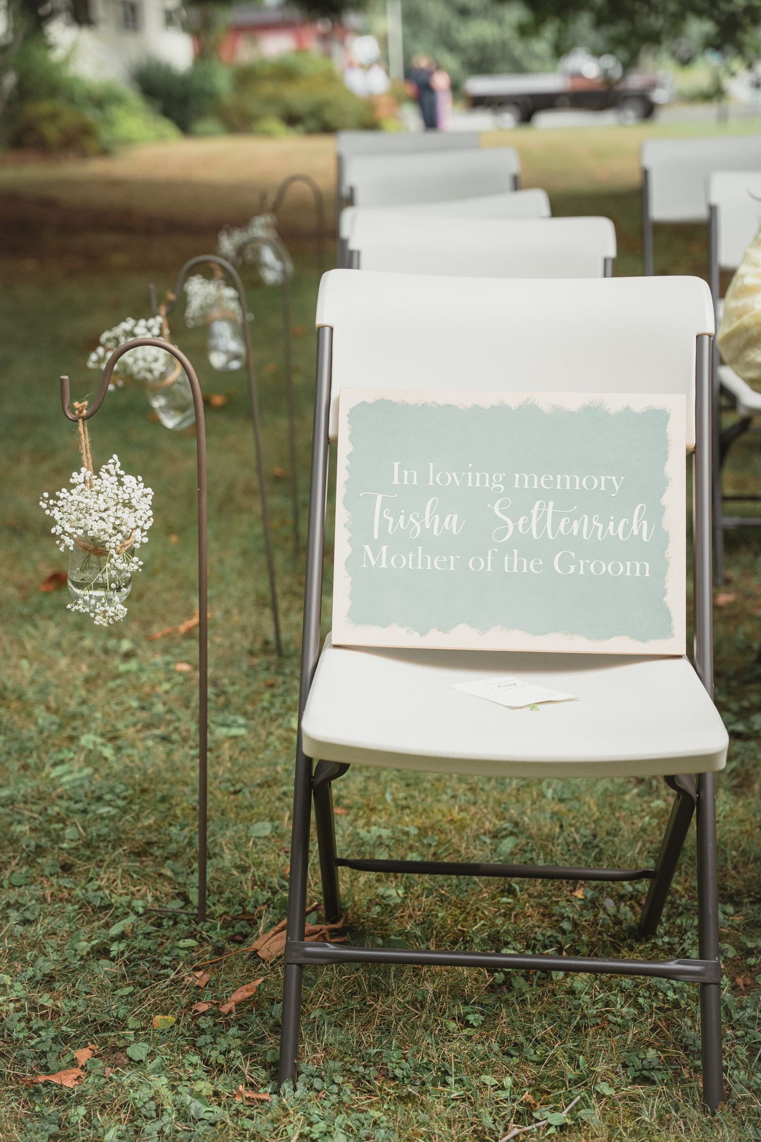 Memorial chair sign reading In Loving Memory Trisha Seltenrich Mother of the Groom at outdoor wedding ceremony.