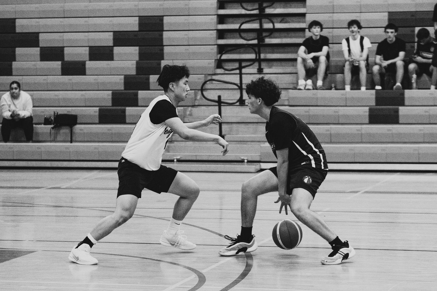 Two young male basketball players compete in a black and white gym during an indoor basketball game.
