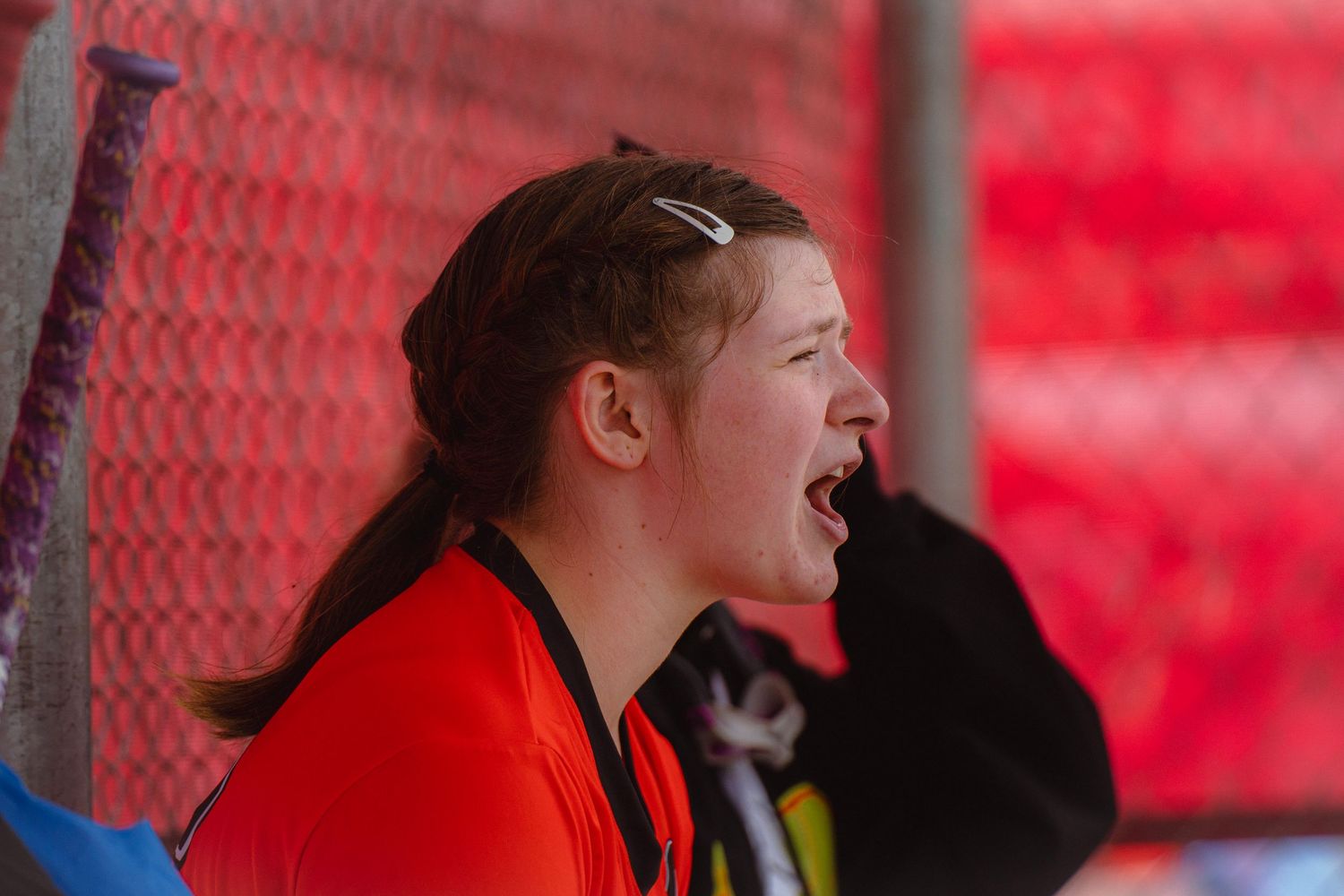 Enthusiastic female softball player in orange jersey cheers loudly from the dugout during a game.