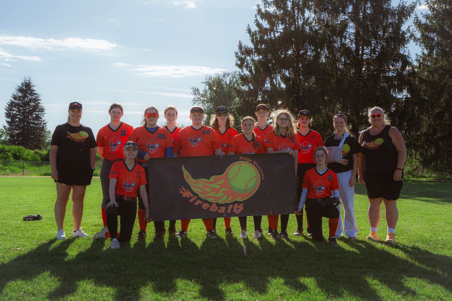 Female softball team in orange jerseys poses outdoors holding a team banner for a group photo.