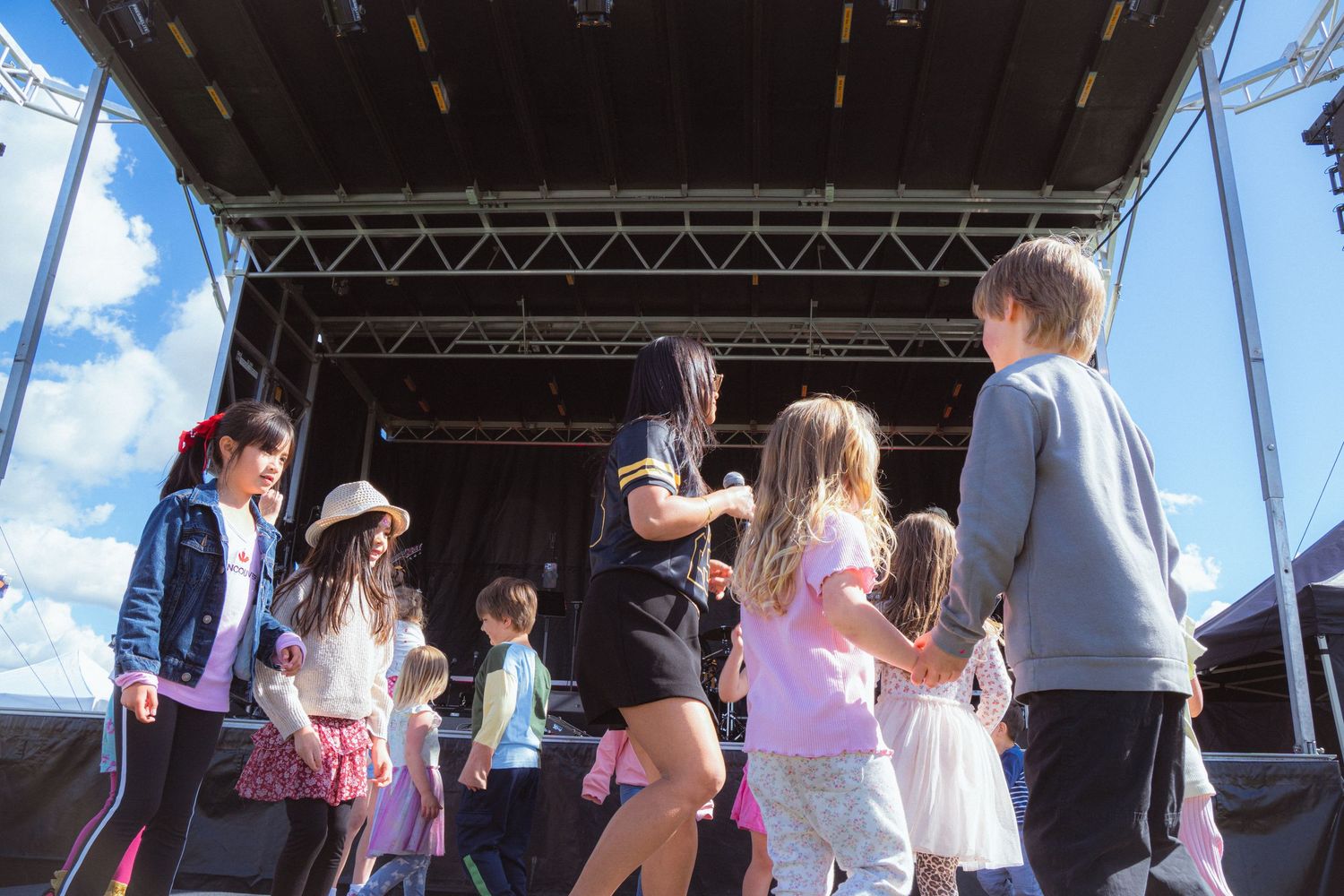 Children and performer dancing together on an outdoor festival stage under a bright blue sky.