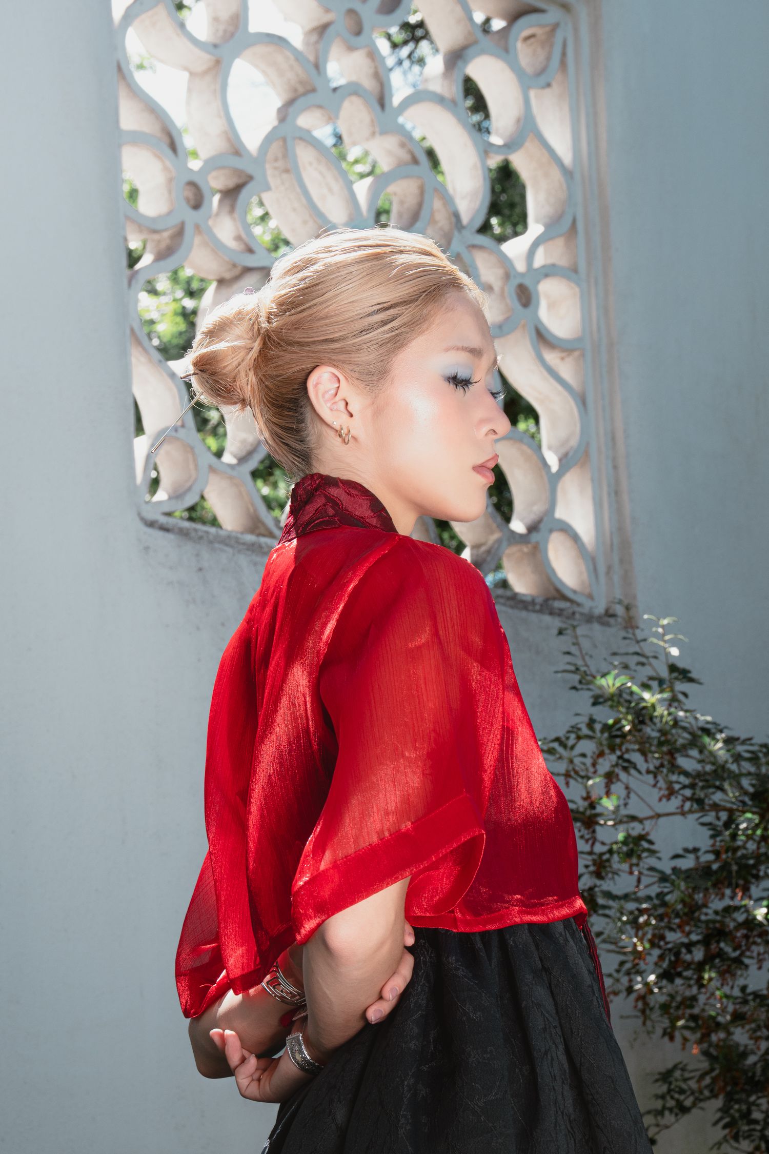 Woman in red sheer kimono top and black skirt posing beside ornate white lattice wall outdoors.