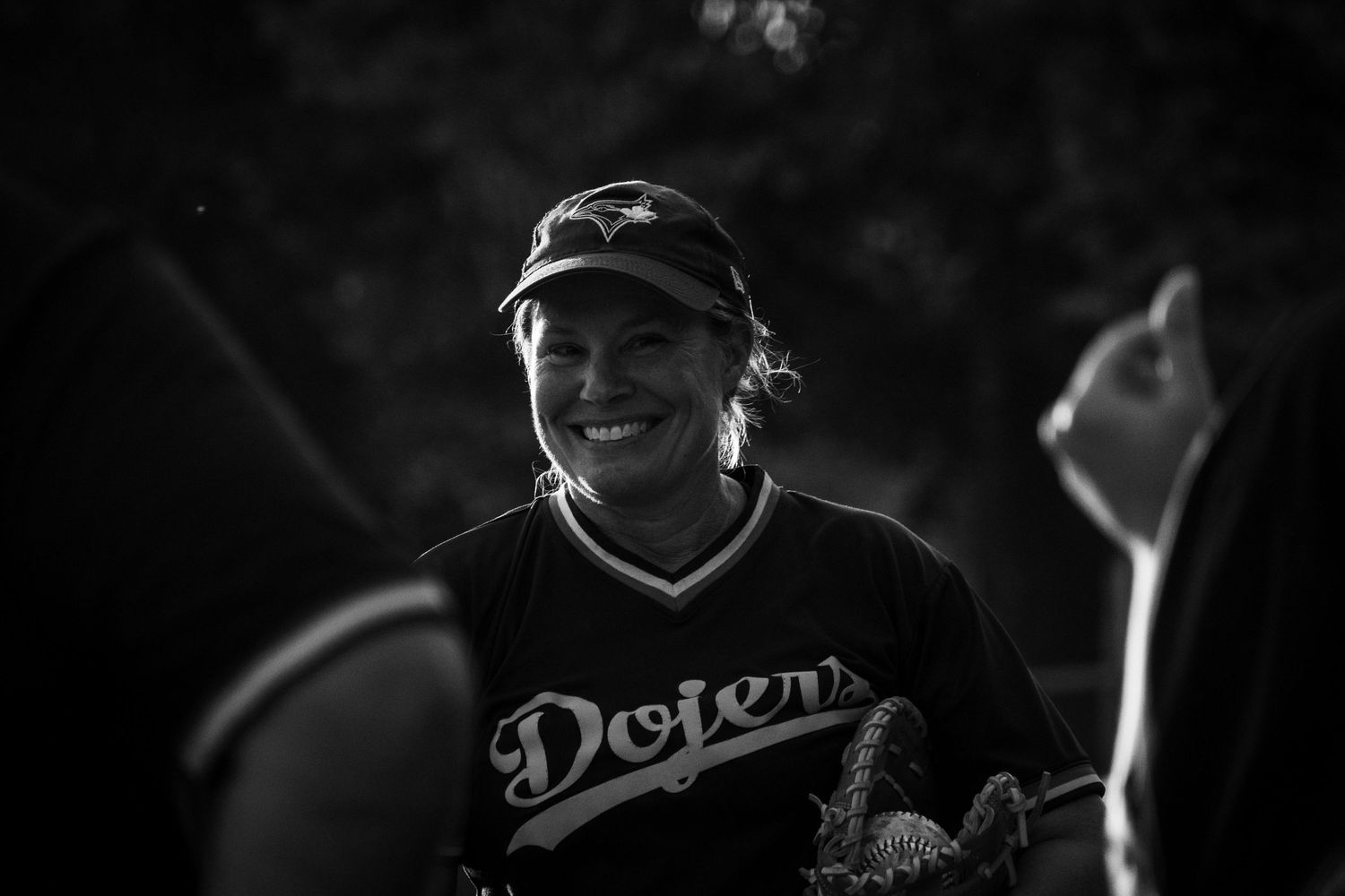 Smiling woman in Dodgers softball jersey and cap photographed in black and white.