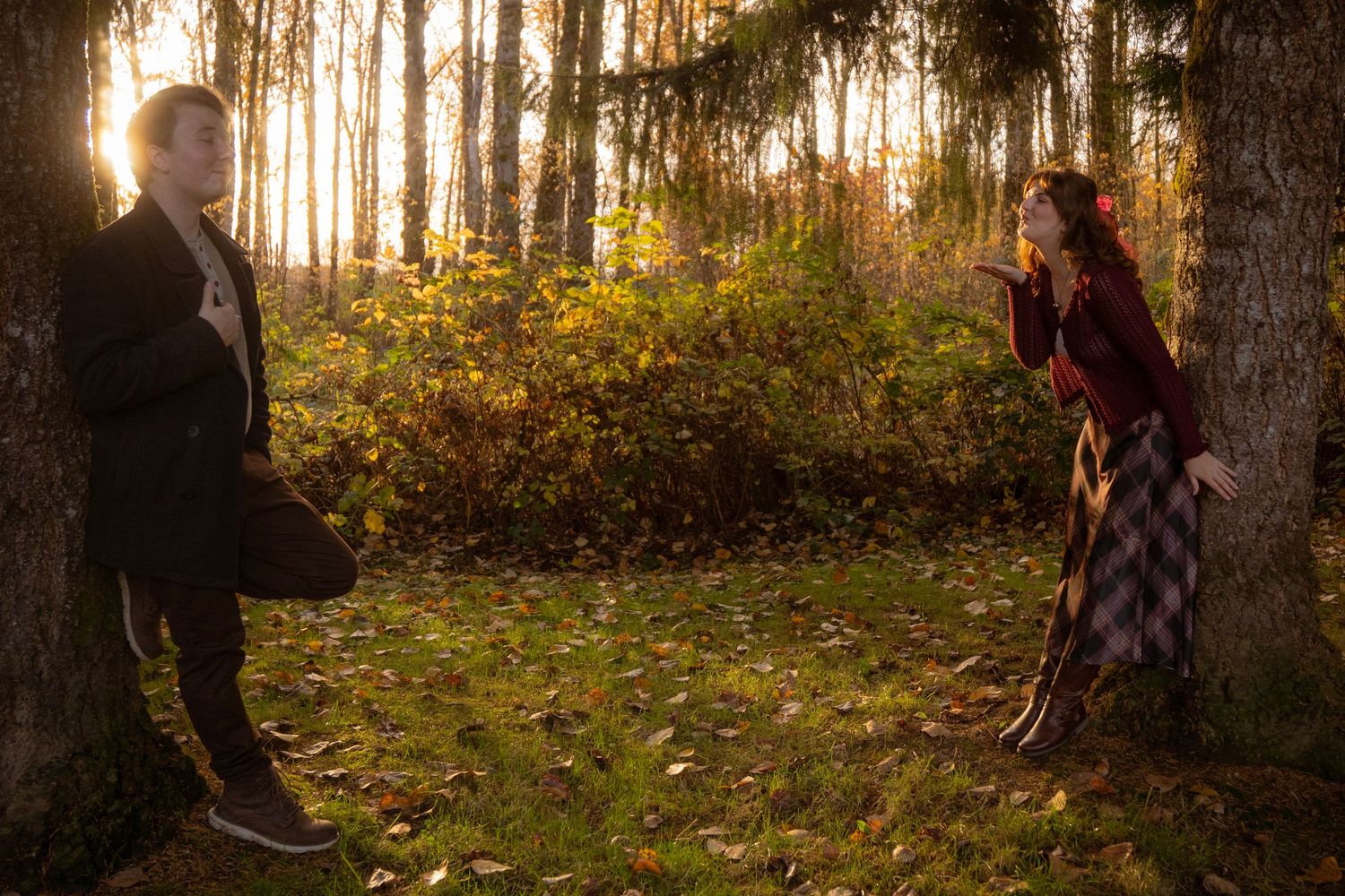 Couple standing apart by trees in golden autumn forest light, facing each other playfully at sunset.