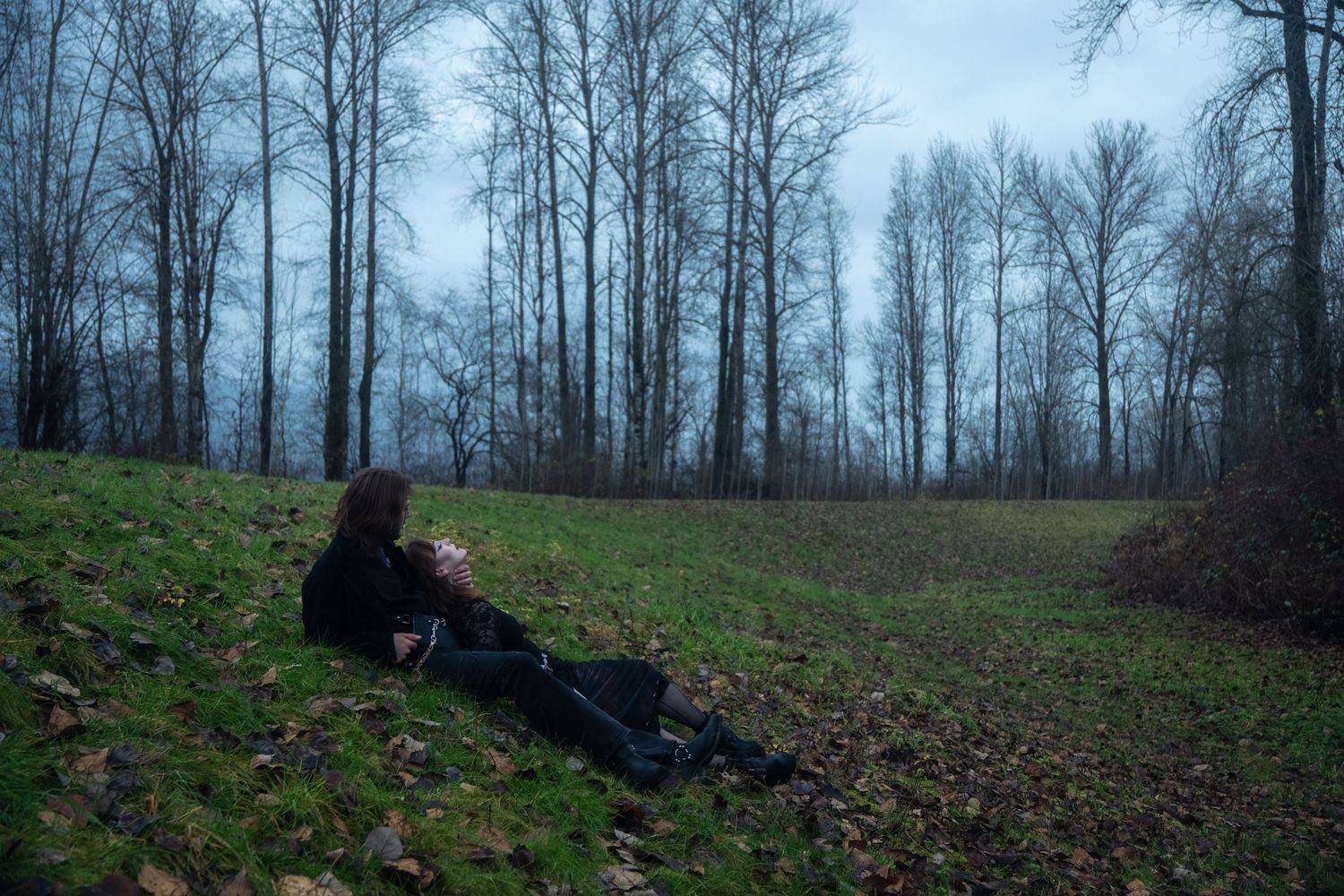 Person in black sitting alone on a grassy slope in a misty bare forest.