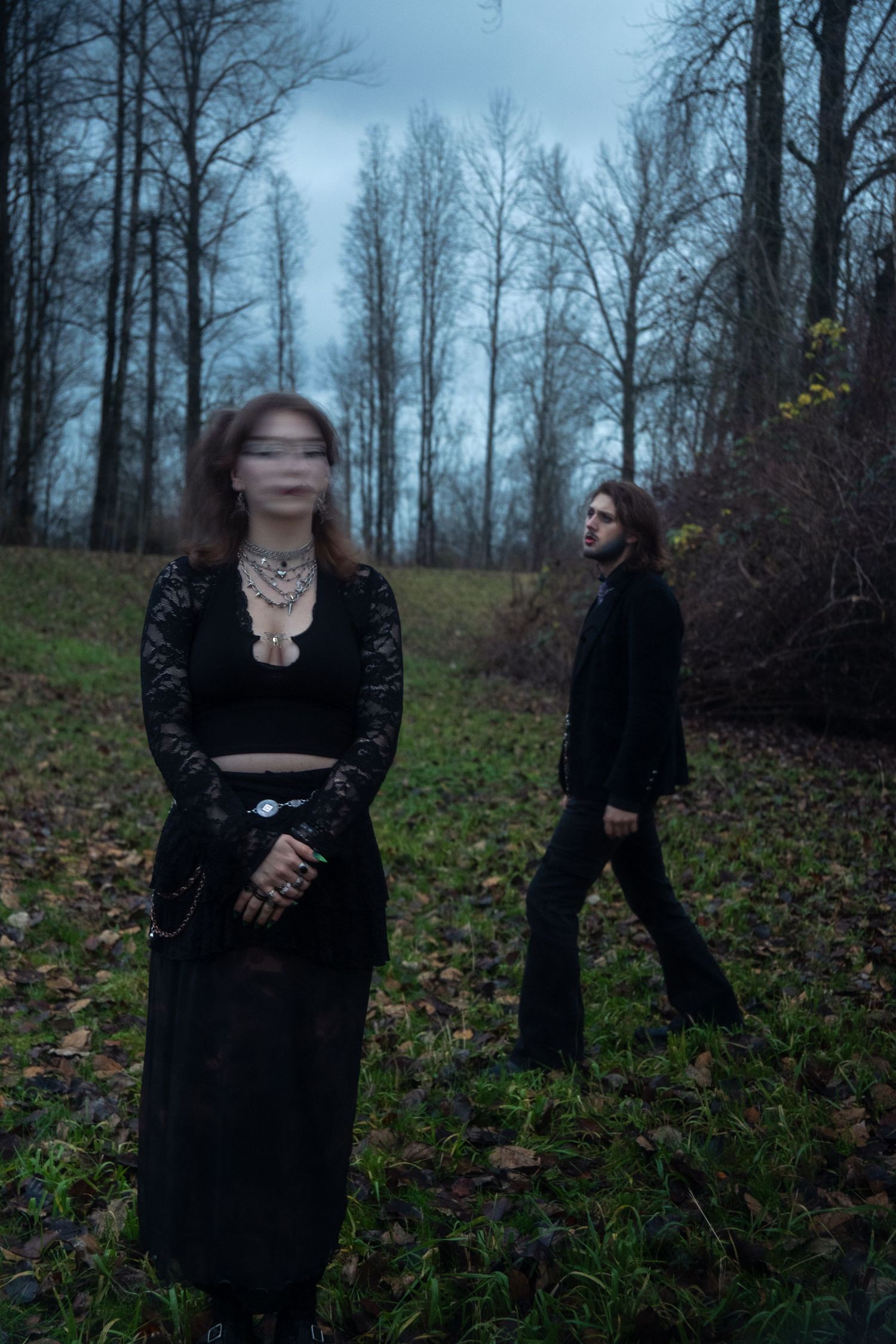 Two gothic figures in black stand in a misty forest clearing with bare trees.