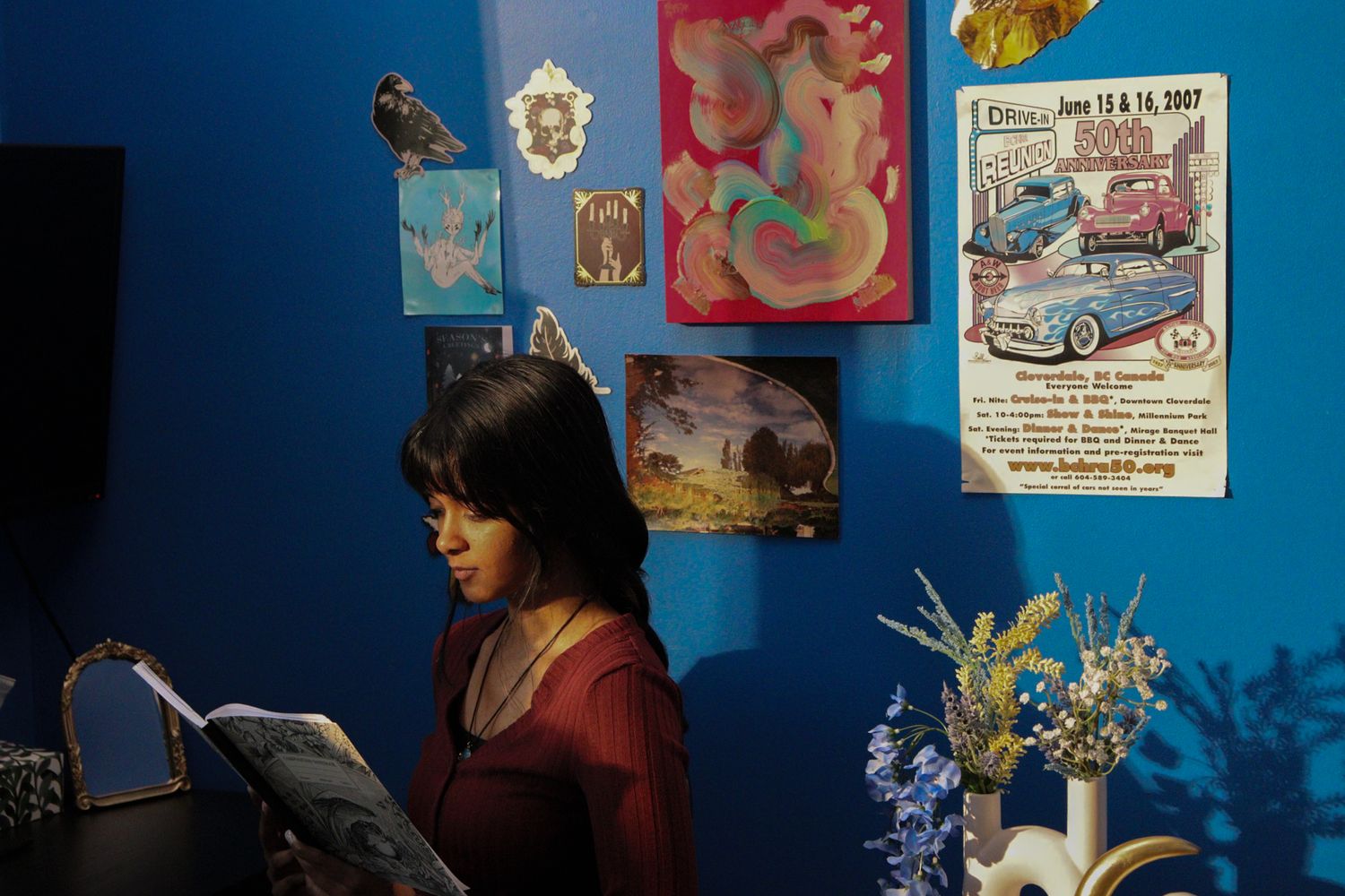 Woman reads book beside colorful posters and floral arrangement on blue wall.