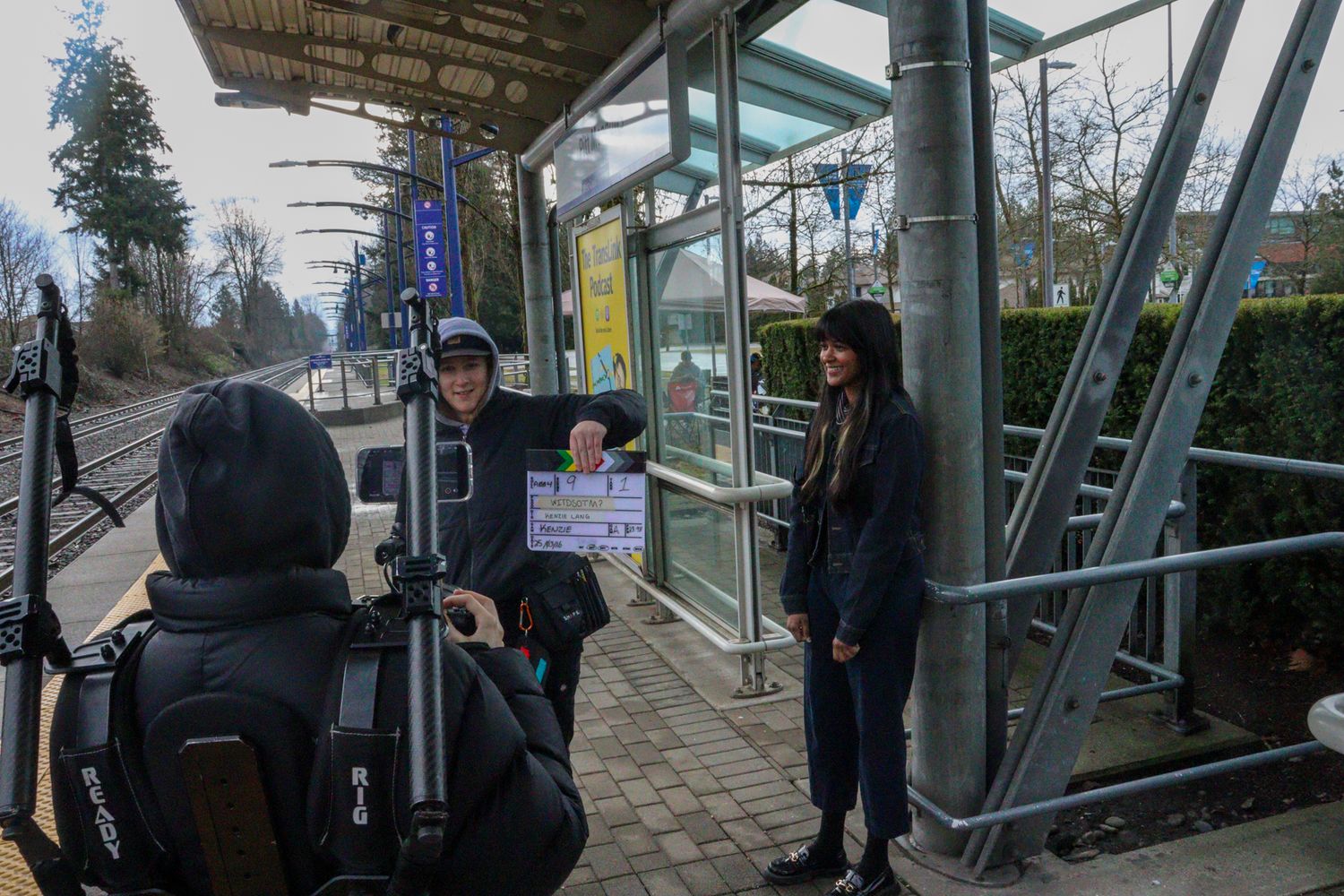 Film crew shoots scene at outdoor transit station with clapperboard and camera.