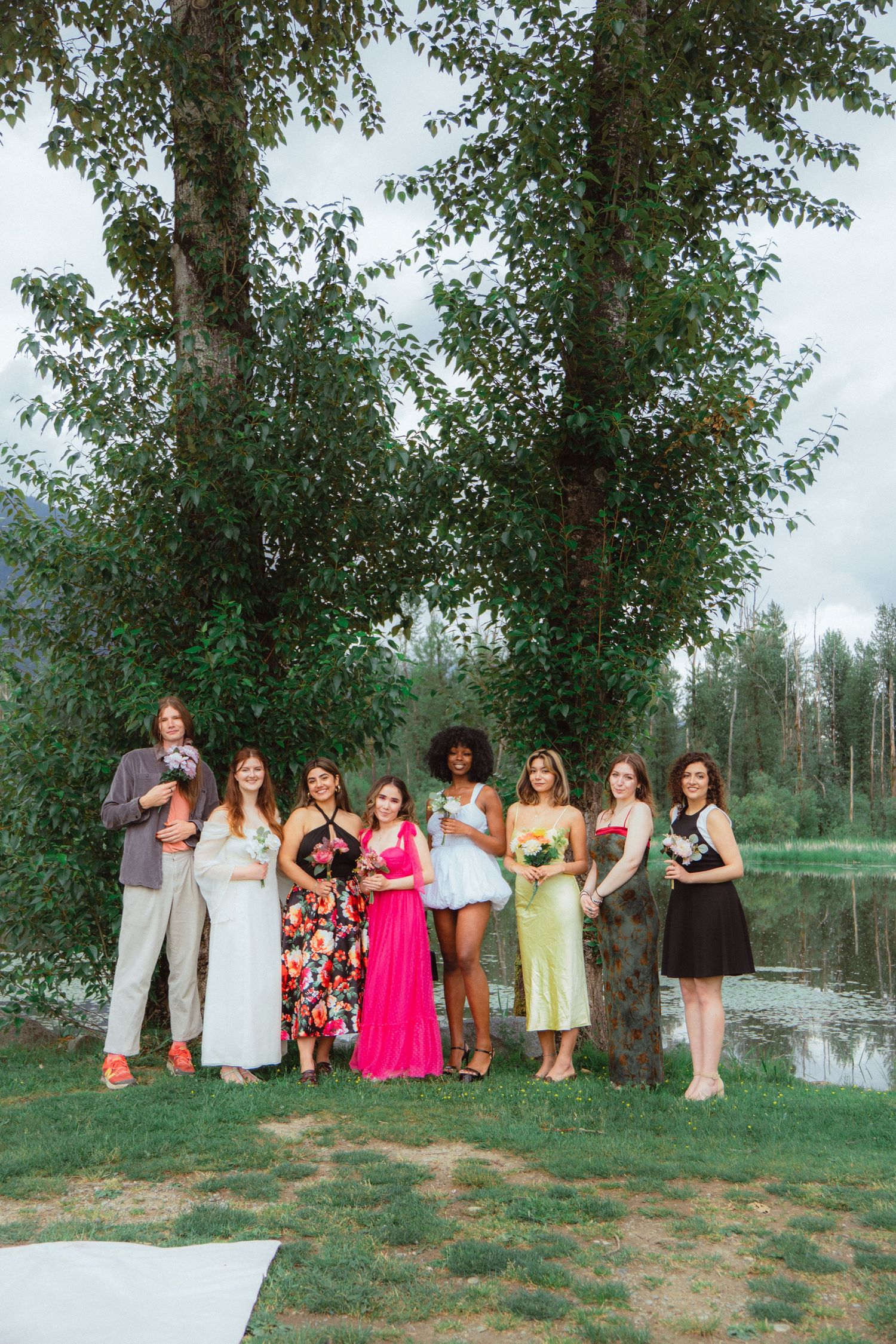 Eight women in colorful dresses standing together holding bouquets near a scenic lake.