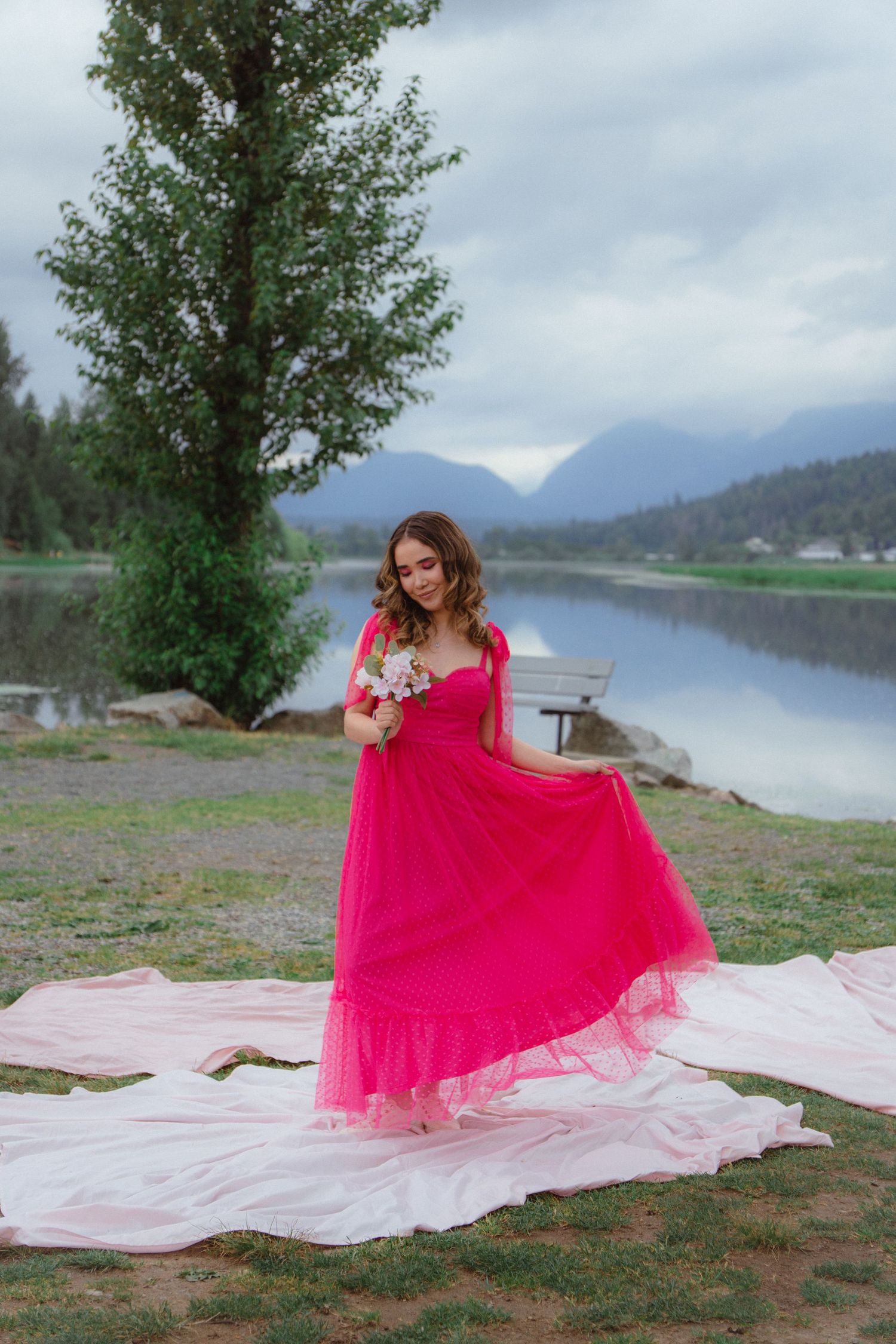 Woman in vibrant pink tulle gown holds flowers by a serene mountain lake on cloudy day.
