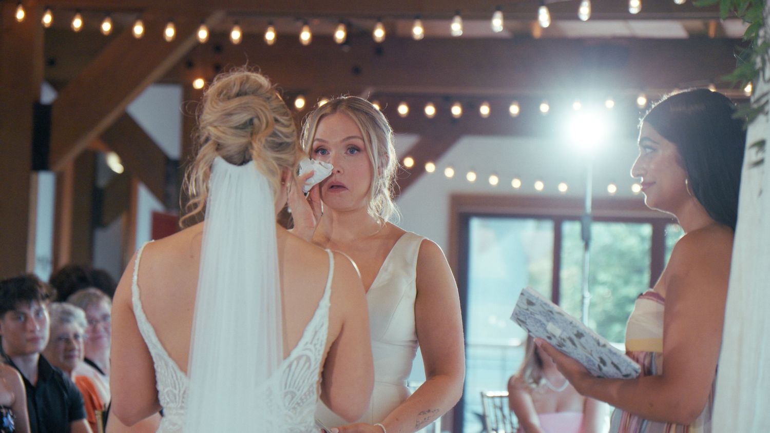 Emotional bride in white gown with veil is comforted by bridesmaid under string lights at ceremony.