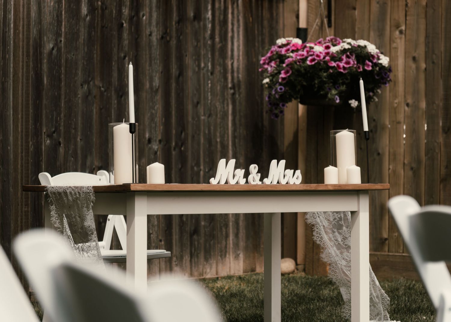 Elegant wedding sweetheart table with Mr & Mrs sign, white candles, and purple floral centerpiece against rustic wood fence.