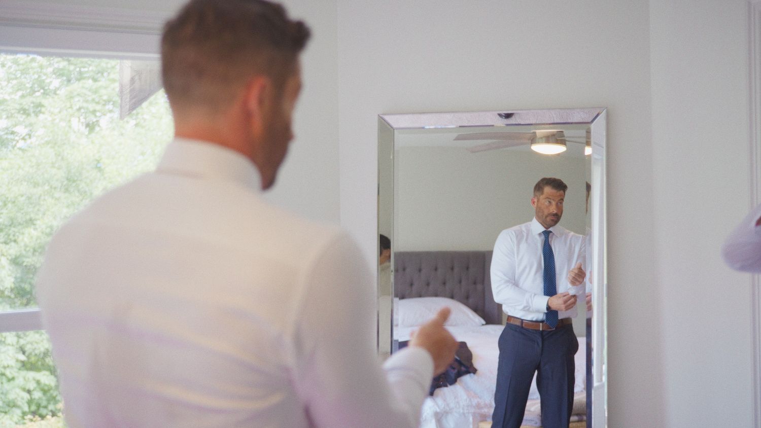 Man in white shirt and tie adjusting cufflinks while looking at his reflection in a bedroom mirror.