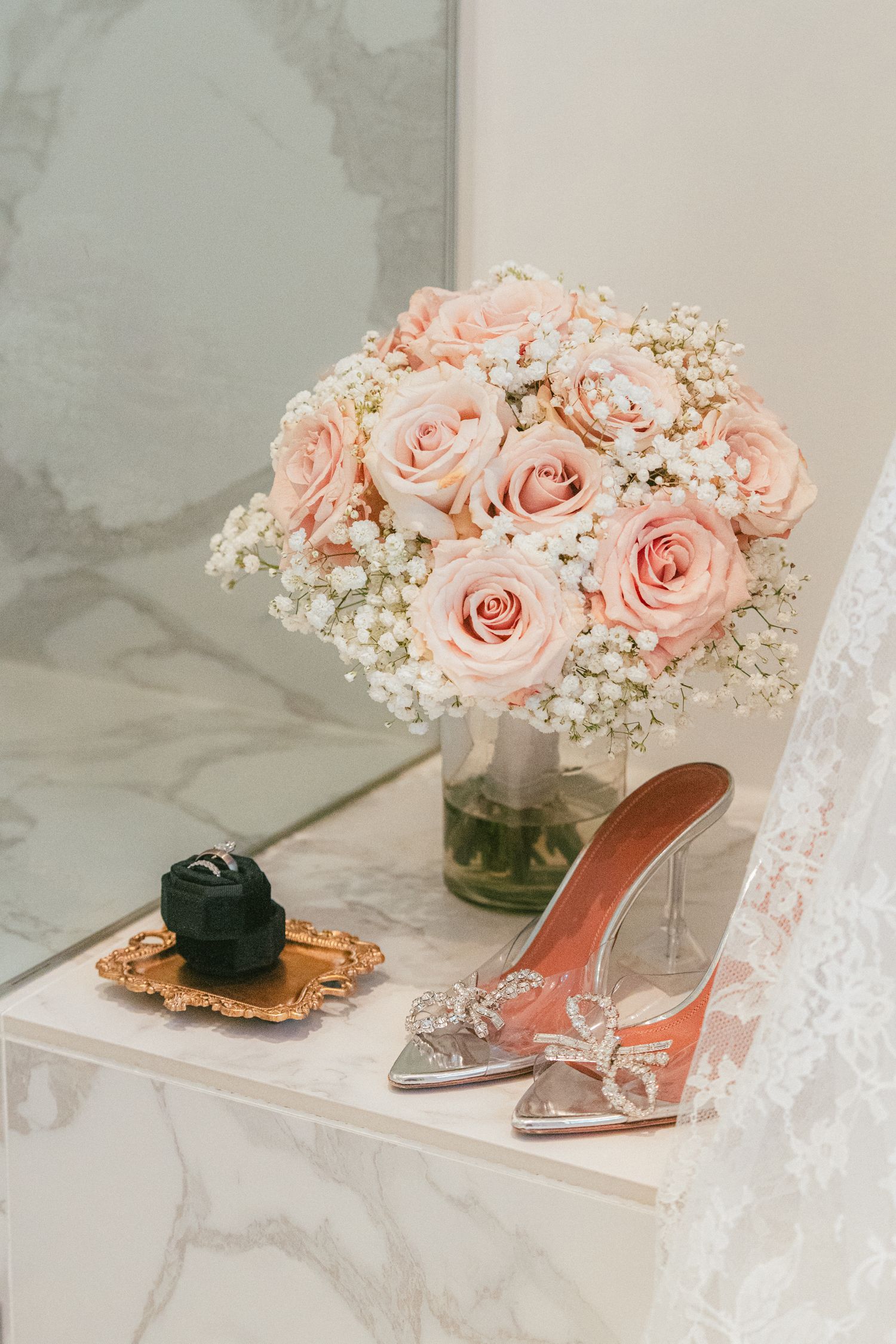 Bridal flat lay with blush pink rose bouquet, crystal heels, and ring box on marble surface.