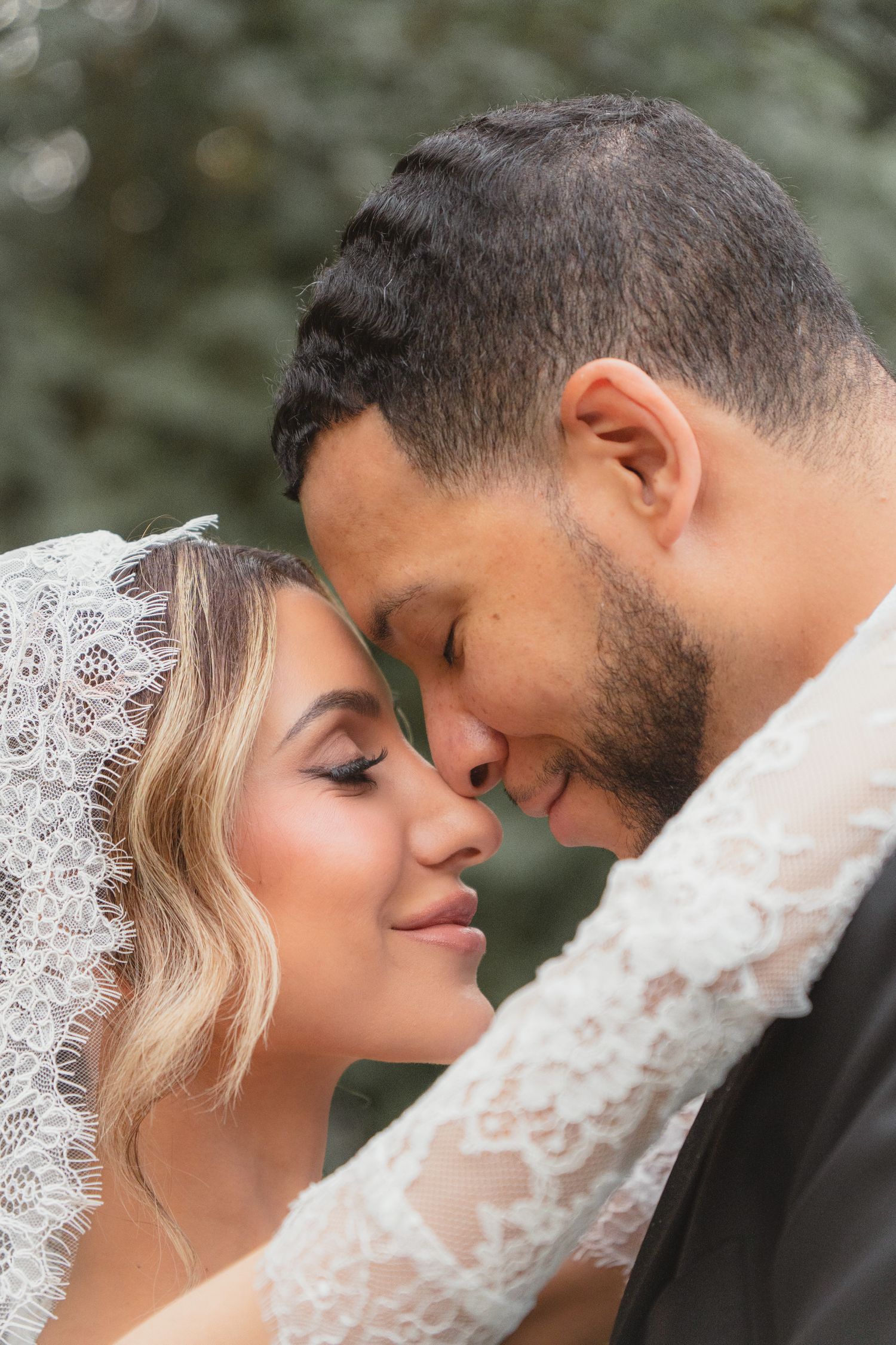 A bride and groom share an intimate forehead-to-forehead moment, with the bride wearing a lace veil on their wedding day.