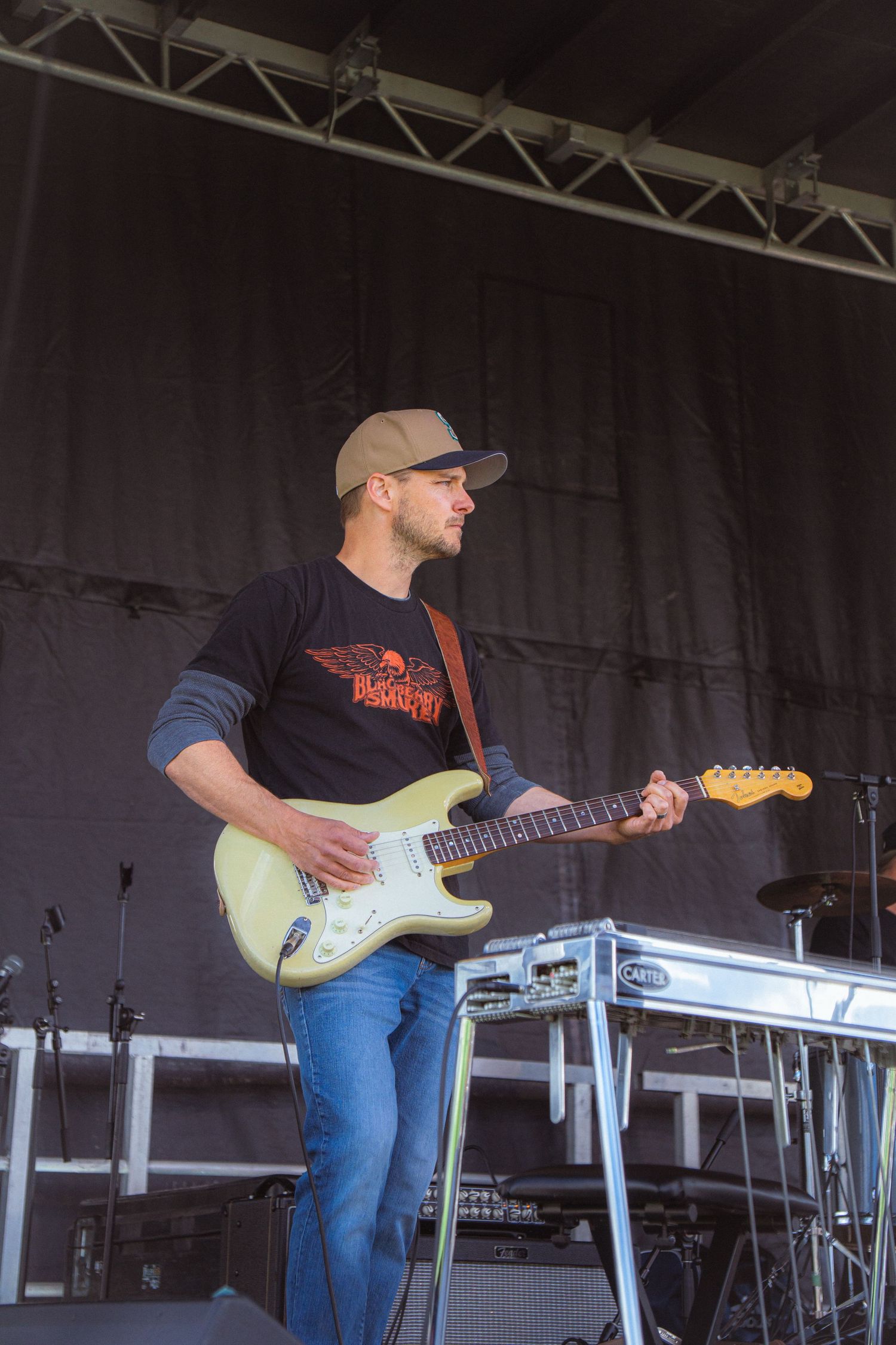 Male guitarist in cap plays white Stratocaster electric guitar on outdoor festival stage.