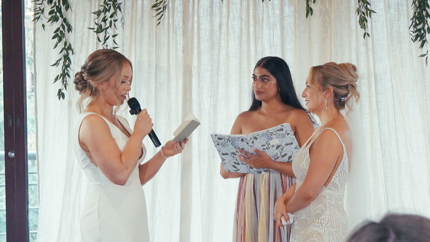 Two brides exchanging vows with officiant at floral altar during intimate wedding ceremony indoors.