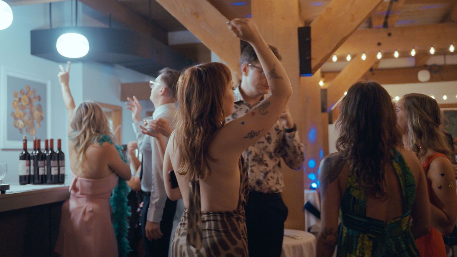 Guests dance and socialize near a bar area with wine bottles at a lively wedding reception.