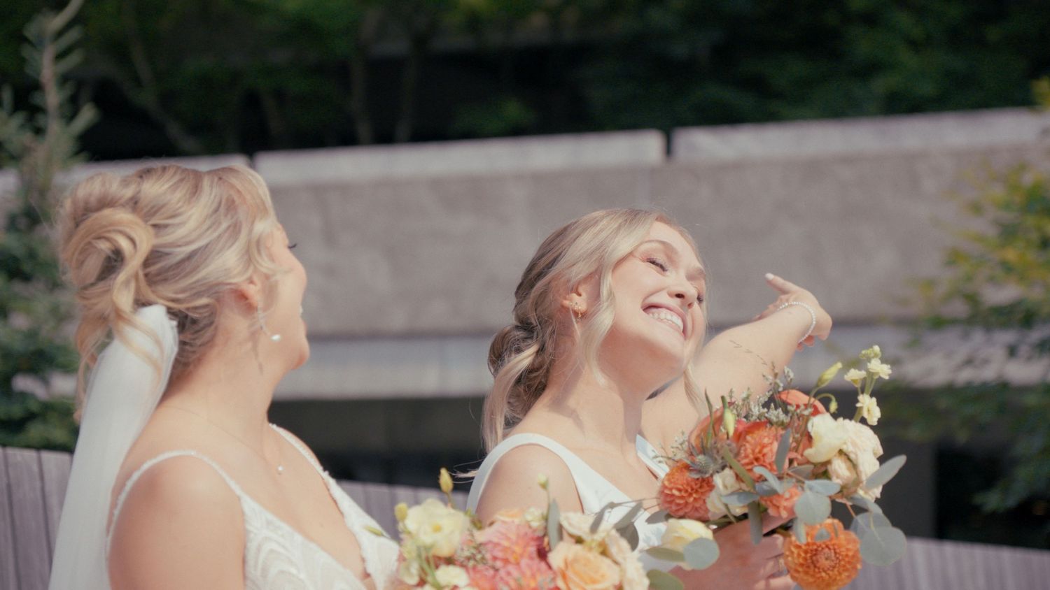 Two brides in white dresses smile and hold colorful bouquets of orange and white flowers outdoors.