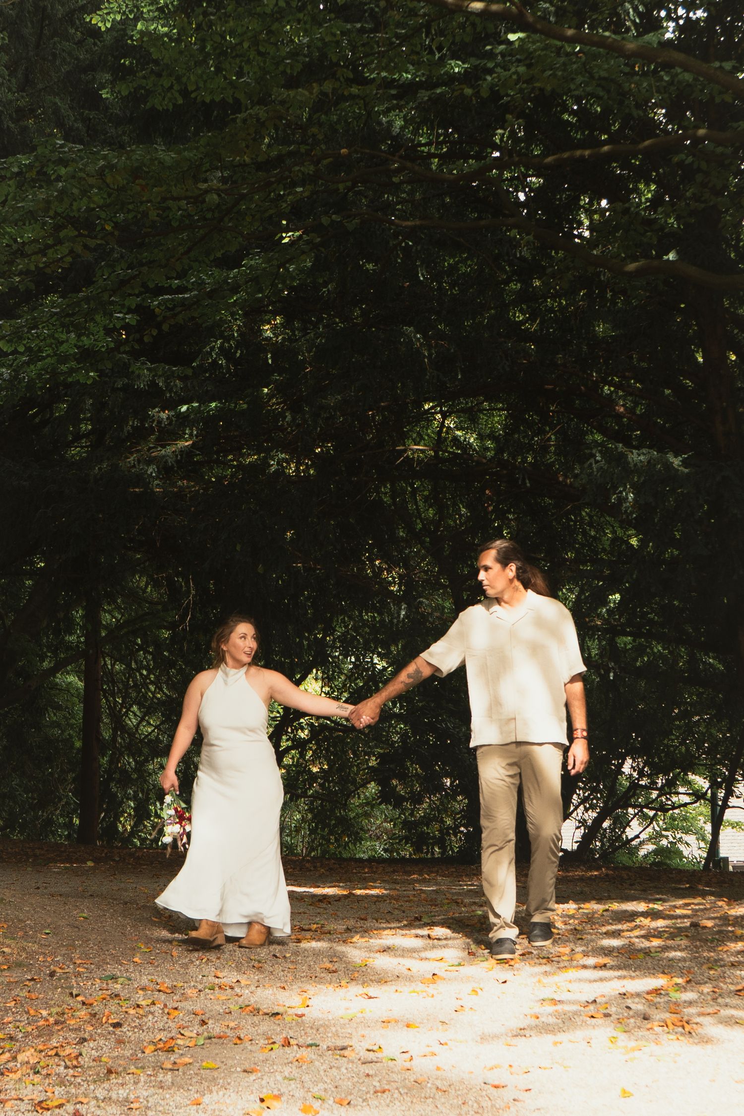 Bride and groom holding hands walking along a sunlit forest path with autumn leaves.