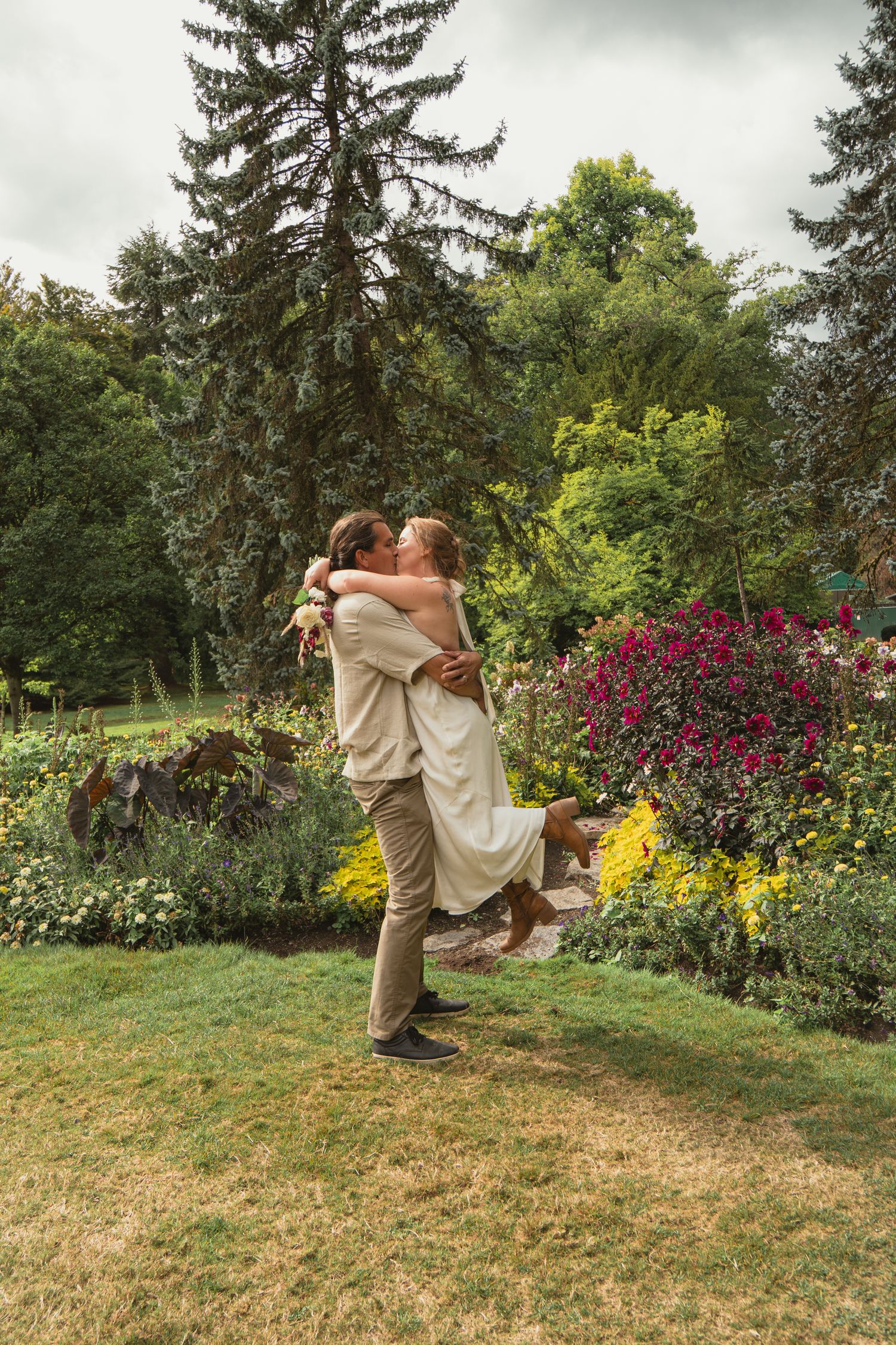Couple sharing a romantic kiss as groom lifts bride in a lush garden with colorful flowers and tall trees.