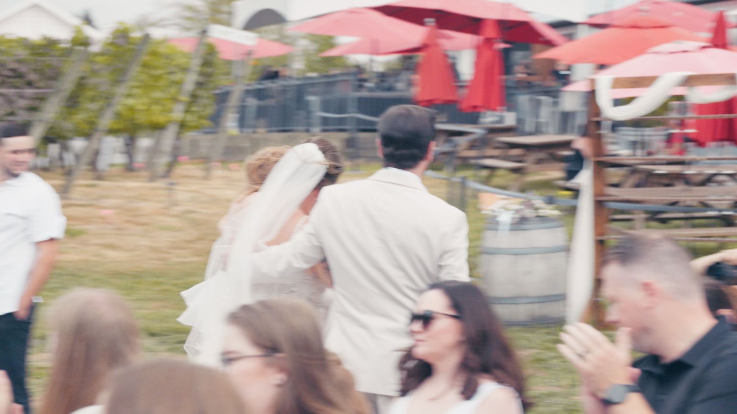 Bride and groom walking together at an outdoor wedding venue with guests and red umbrellas in the background.