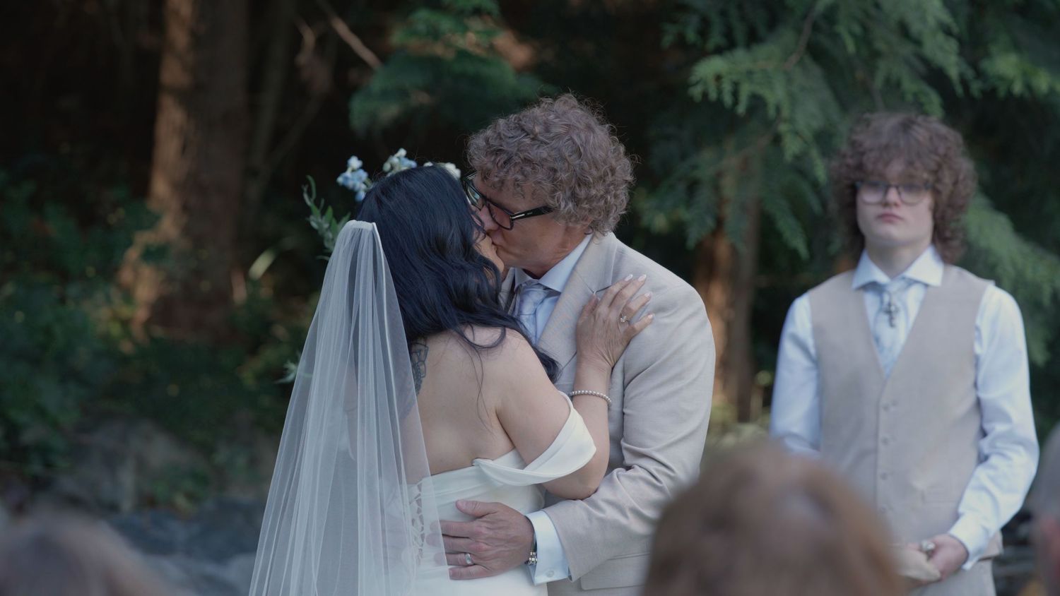 Bride and groom share first kiss at outdoor wedding ceremony while guests watch.
