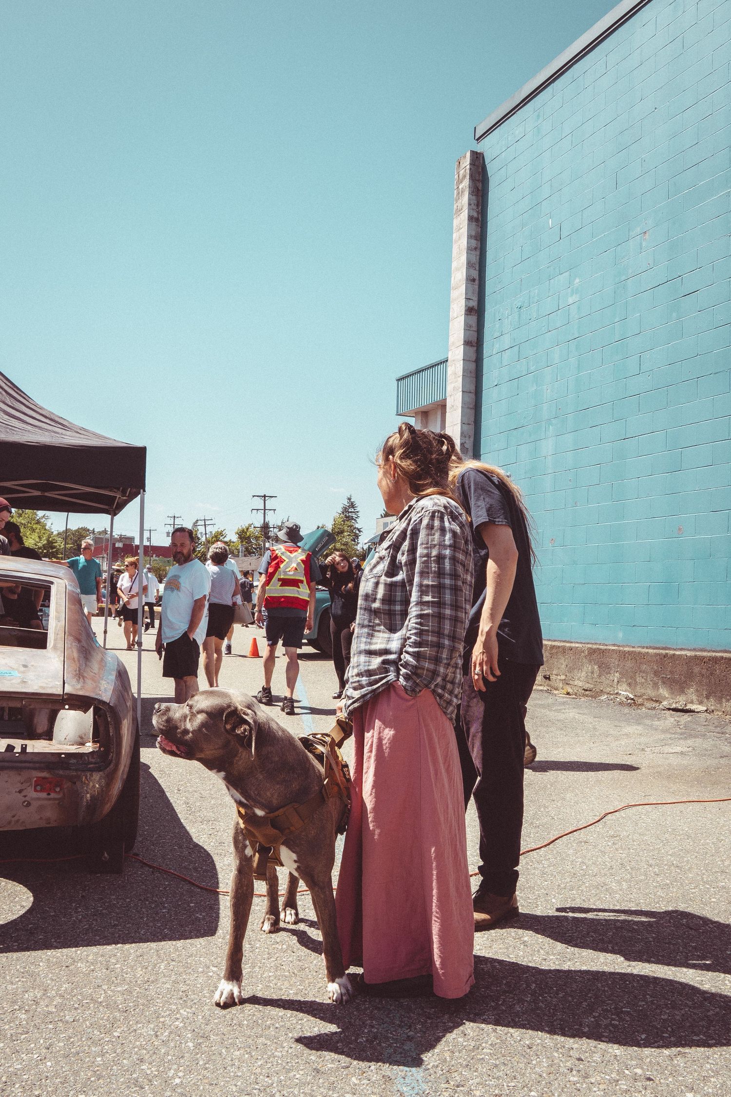 Woman in pink skirt with brindle dog on leash at a sunny outdoor car show near a blue building.