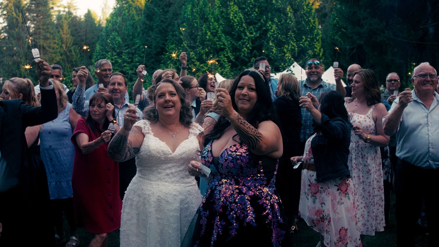 Two women celebrating at an outdoor wedding surrounded by cheering guests at dusk.