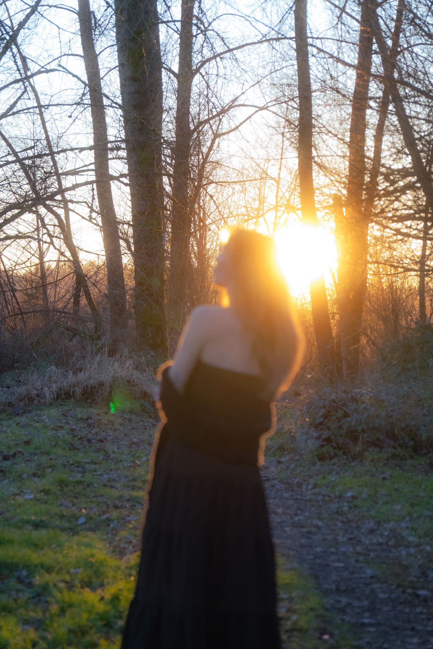 Blurry silhouette of a woman in a black dress standing in a forest path at golden sunset.