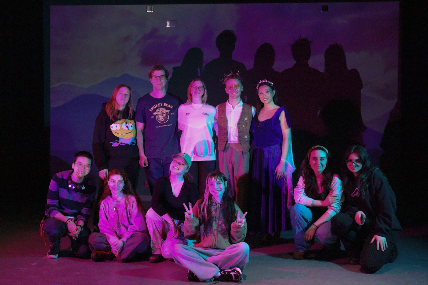 Group of students pose together on stage under purple and pink lighting with projected shadows behind them.