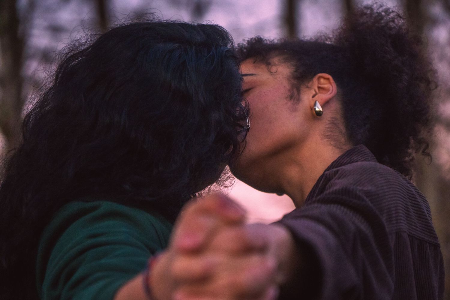 Two women share an intimate kiss while holding hands outdoors during a moody purple twilight evening.