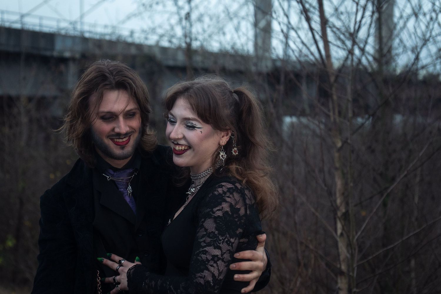 Smiling gothic couple in black embrace outdoors near bare trees and bridge structure.