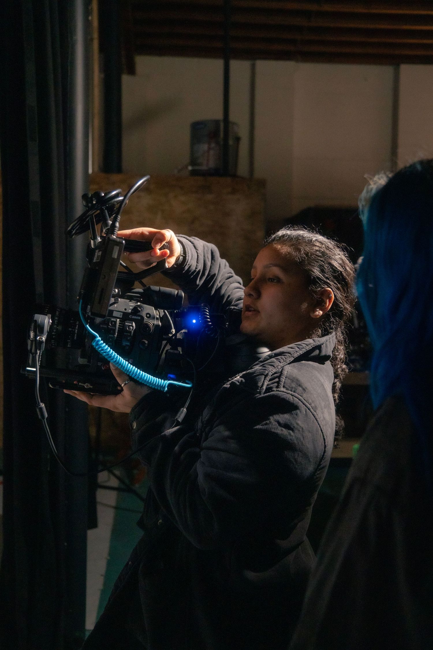 A filmmaker adjusts a professional video camera with a blue coiled cable in a dimly lit studio setting.