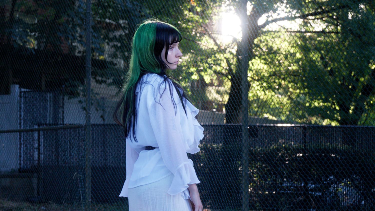 Woman with green hair in white outfit stands in profile by chain-link fence in sunlight.