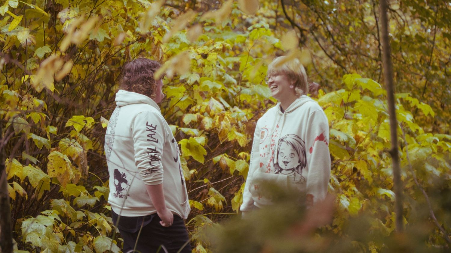 Two people wearing matching white graphic hoodies stand facing each other among autumn yellow foliage.