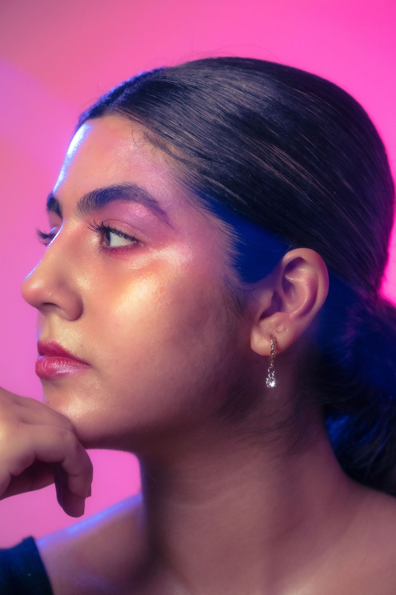 Side profile of young woman with pink eyeshadow and dangling earring resting chin on hand in neon pink light.