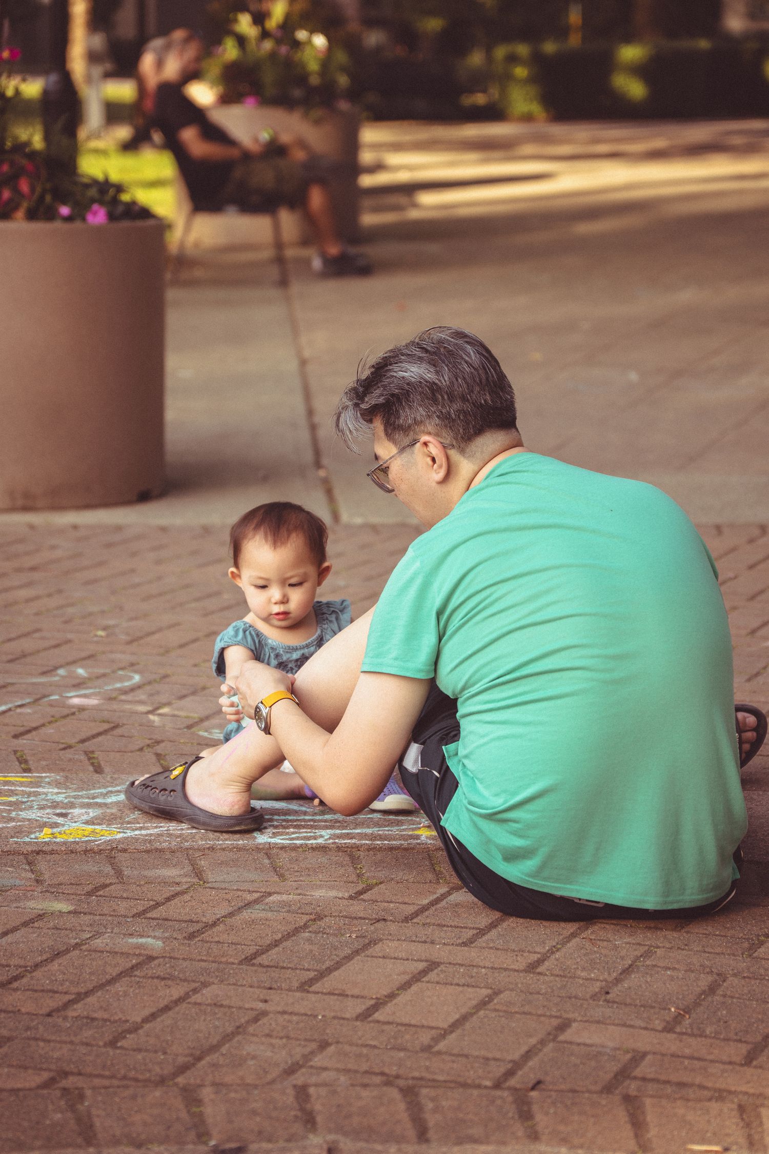 Father and toddler drawing with chalk on brick pavement in a sunny outdoor park setting.
