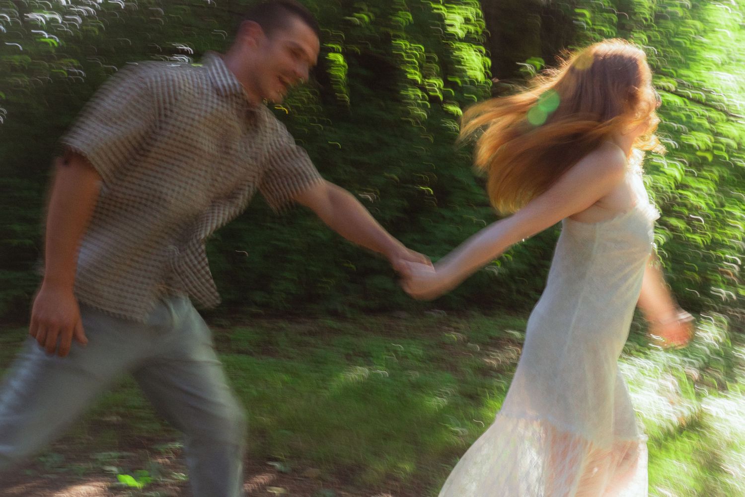 Couple holding hands and spinning joyfully in a sunlit forest, captured with motion blur effect.