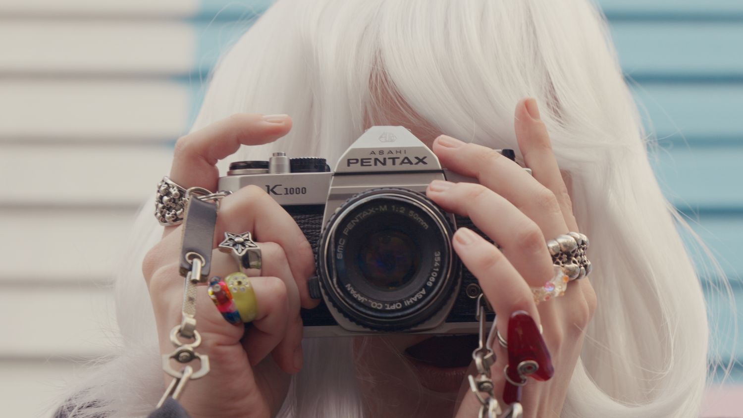 Blonde woman holding a Pentax film camera up to her face, wearing multiple rings on her fingers.